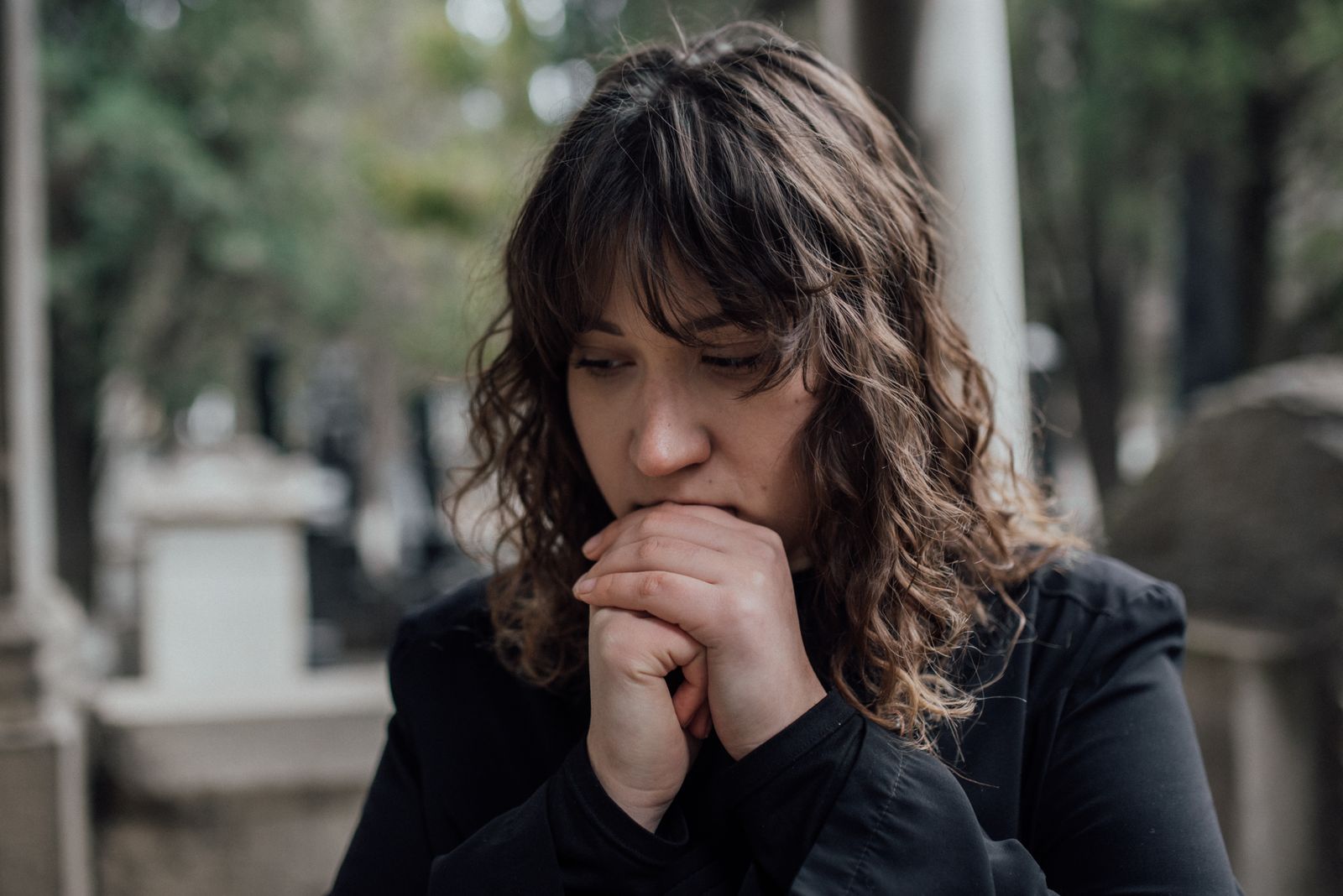 Pensive woman in black coat standing outdoors with hands together, conveying sadness.