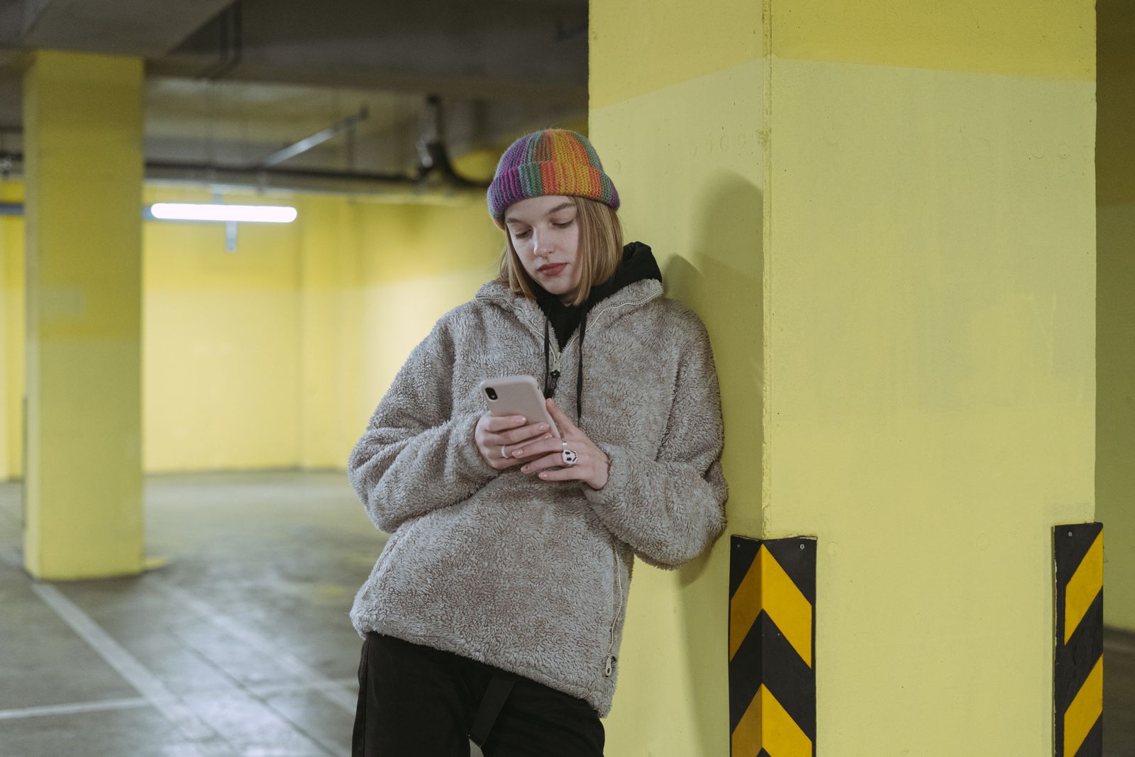 Woman in colorful beanie leaning on a yellow pillar, texting on her phone in an indoor parking garage.