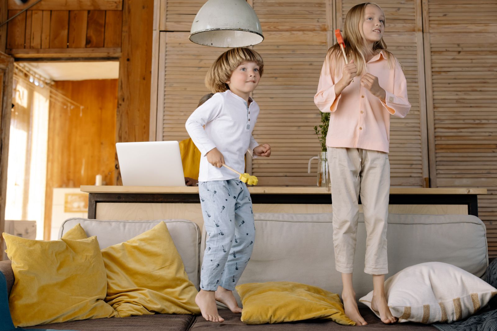 Two kids having fun playing and jumping on a cozy sofa indoors with pillows scattered around.