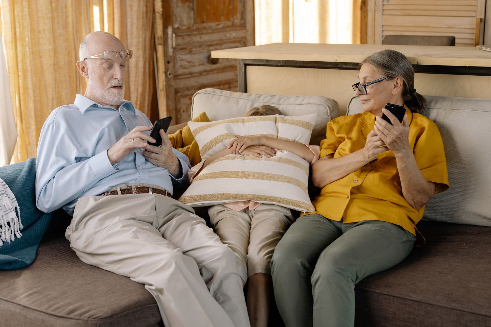 Senior couple on sofa with grandchild, using smartphones in cozy indoor setting.
