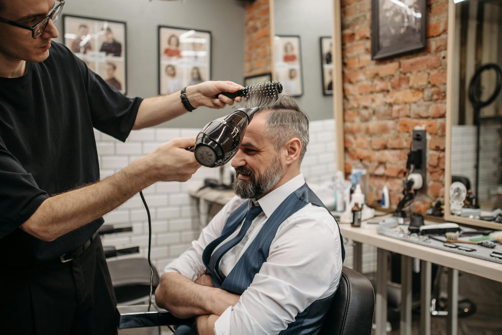 An elderly man gets his hair blow-dried by a barber in a modern barbershop setting.