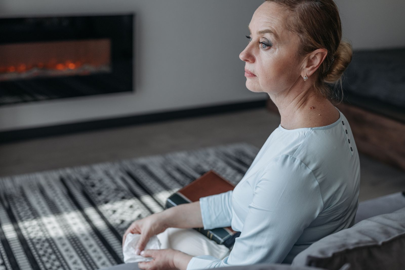 Senior woman sitting indoors, reflecting near a warm fireplace, calm atmosphere.