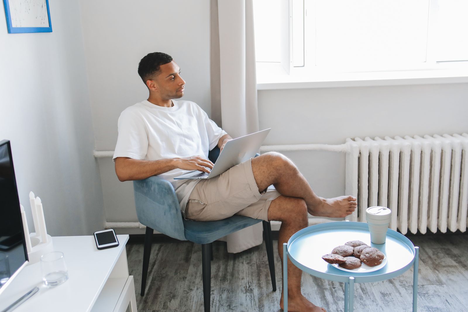 Casual home setting with man using laptop, enjoying cookies and coffee by a window.