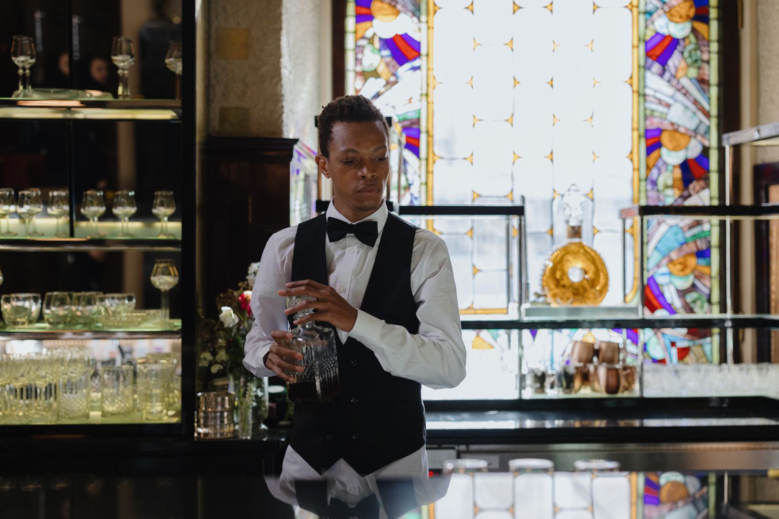 Bartender skillfully handling cocktails behind the counter in a classic, upscale bar setting with stained glass backdrop.