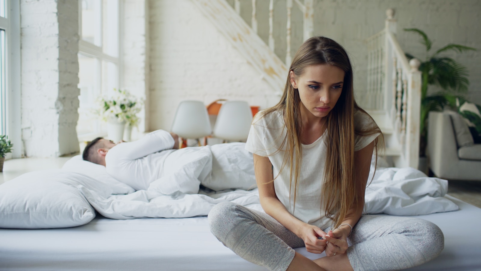 Woman sitting on bed, man sleeping behind her.