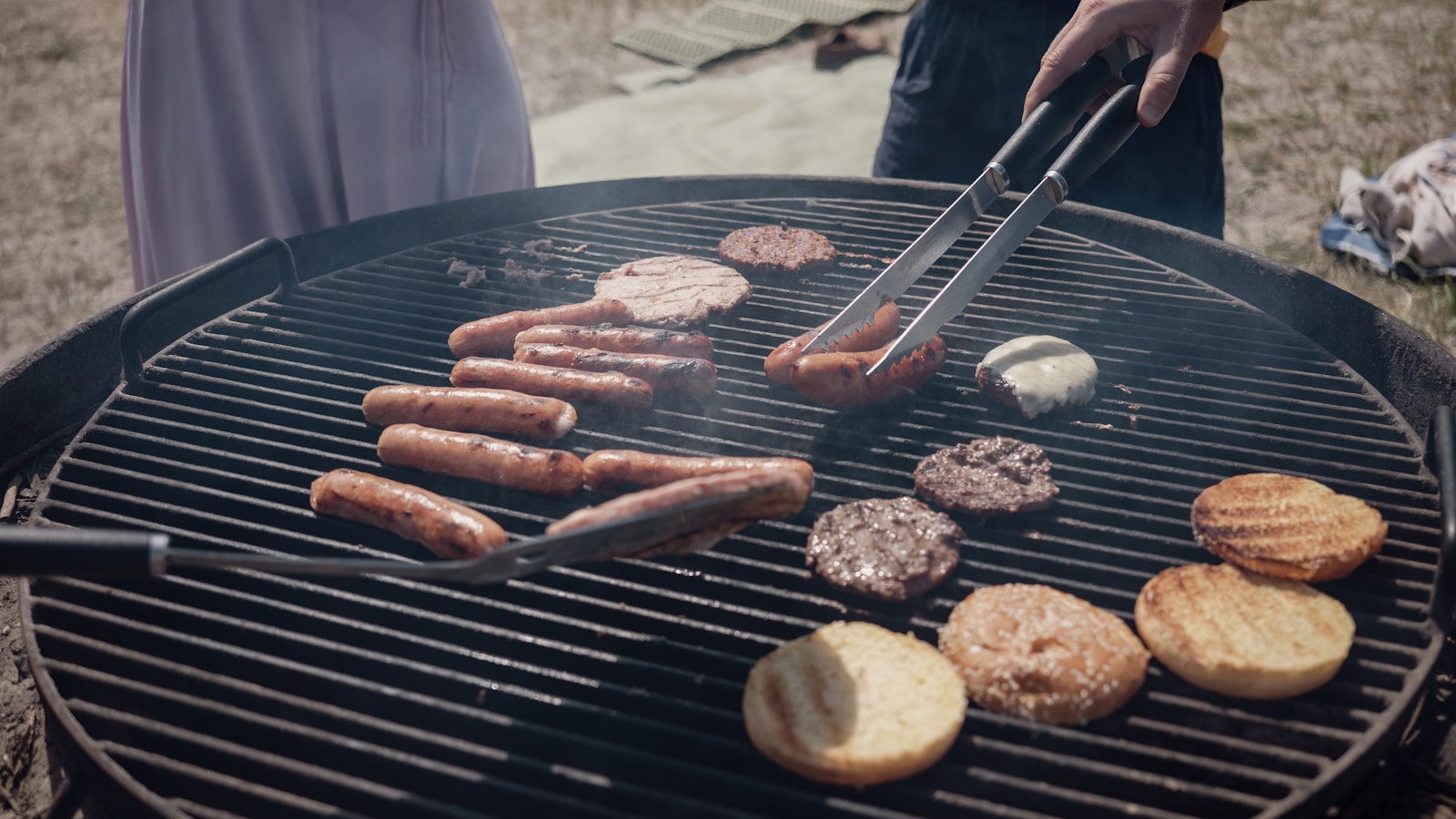 Hand turning sausages and burgers on an outdoor grill.
