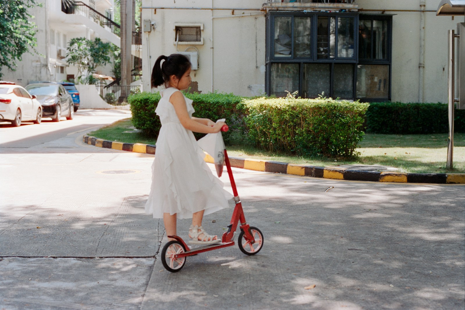 Young girl in white dress rides red scooter outdoors