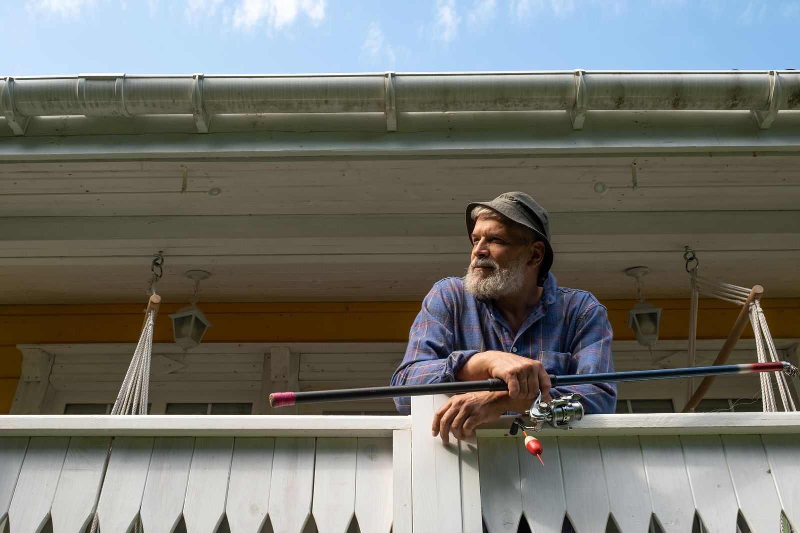 A senior man leans on a porch railing holding a fishing rod, enjoying the outdoors.
