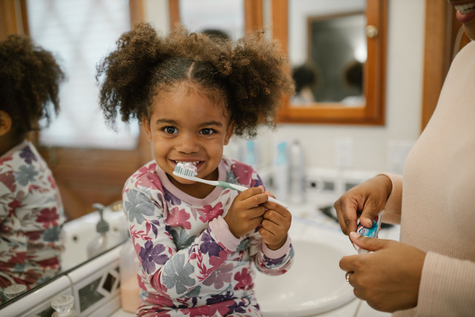 Young girl with curly hair brushing teeth supervised by parent in a home bathroom.