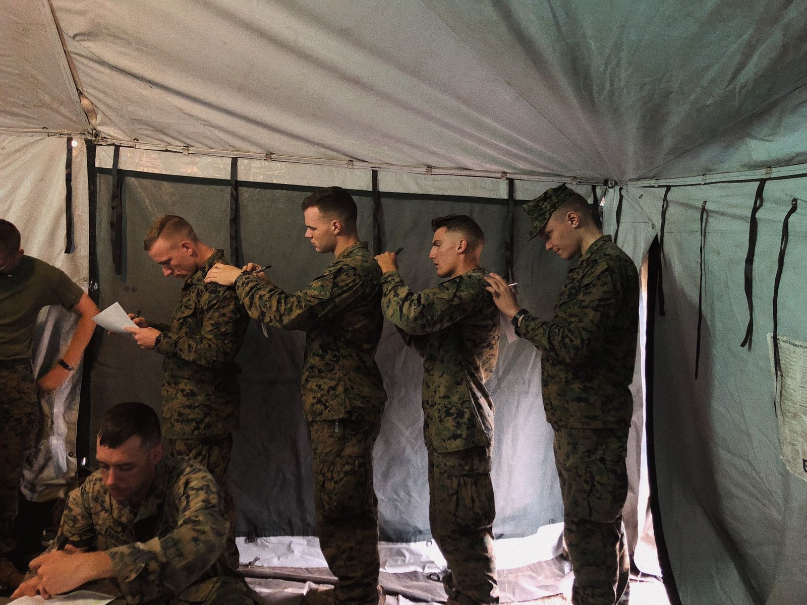 Soldiers writing on each other's backs inside a camouflage military tent.