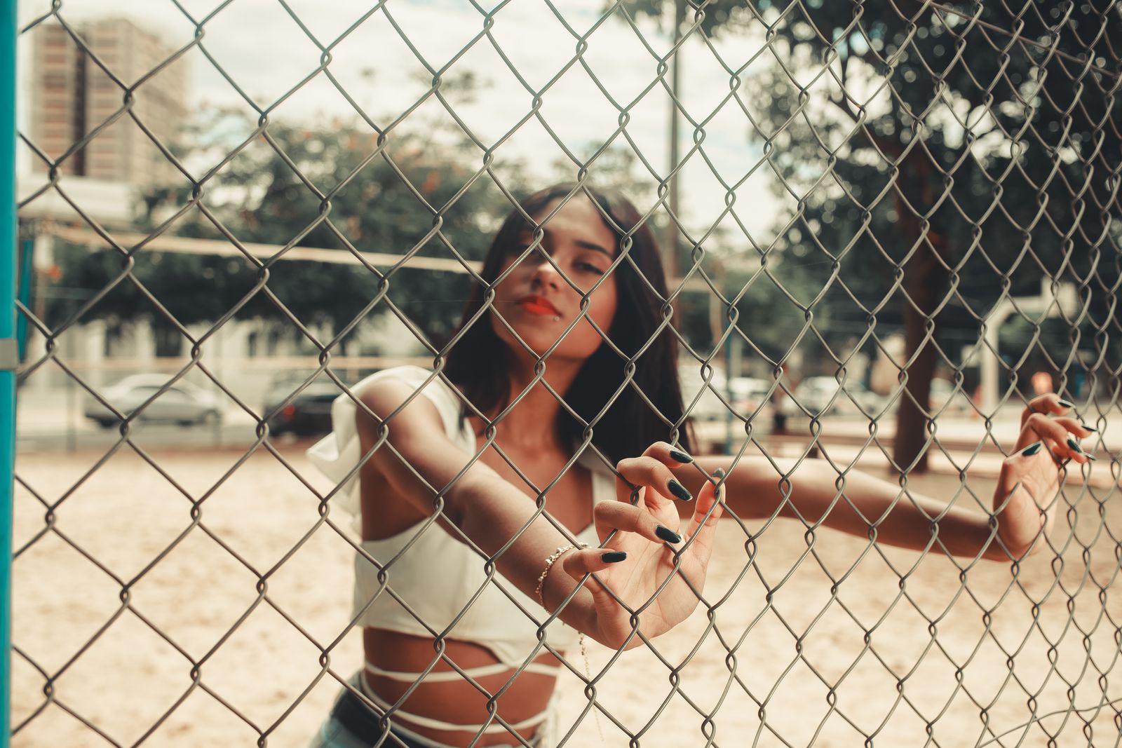 Stylish young woman in summer attire poses behind wire mesh fence outdoors.