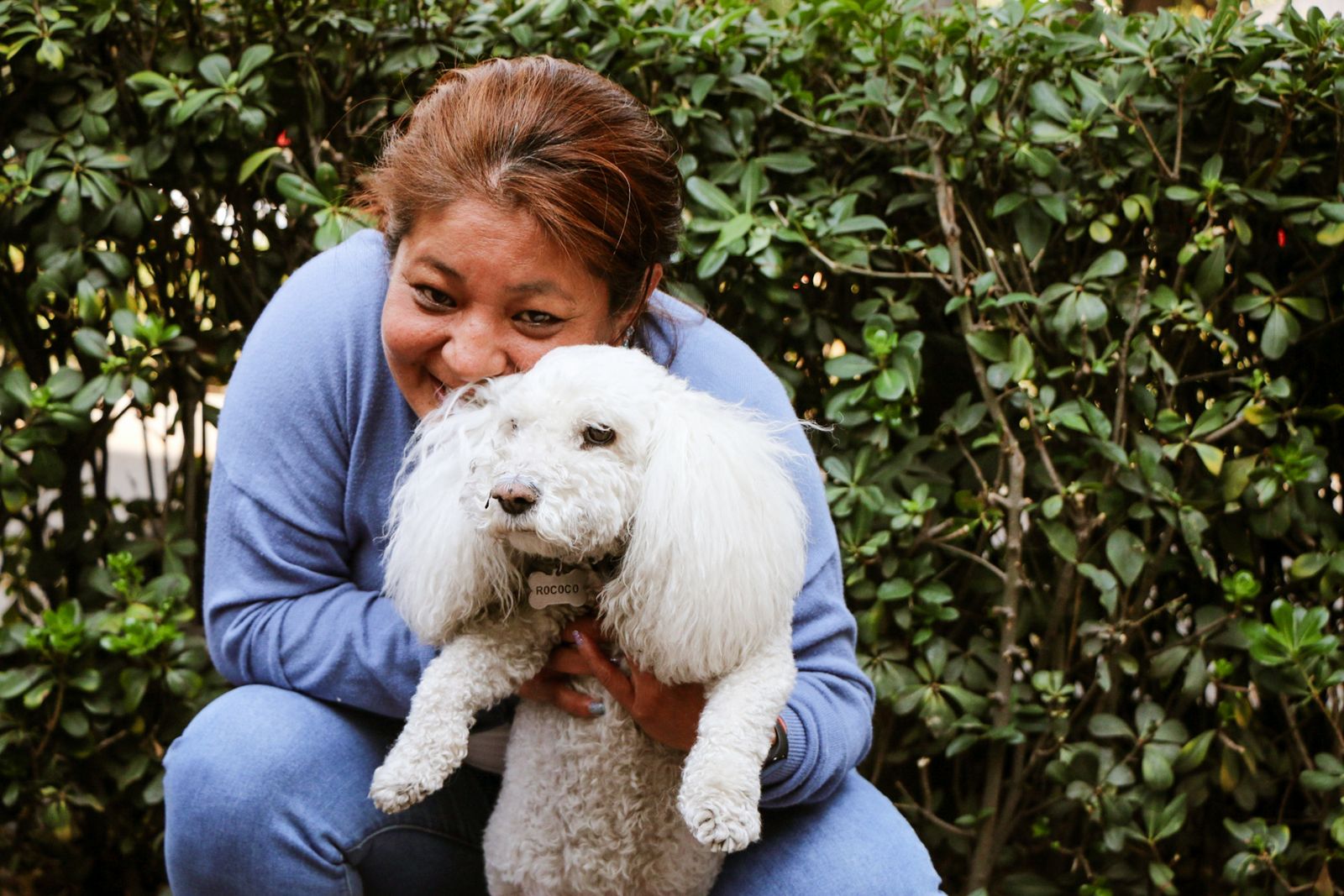 A woman lovingly hugs her white poodle outdoors with greenery in the background.