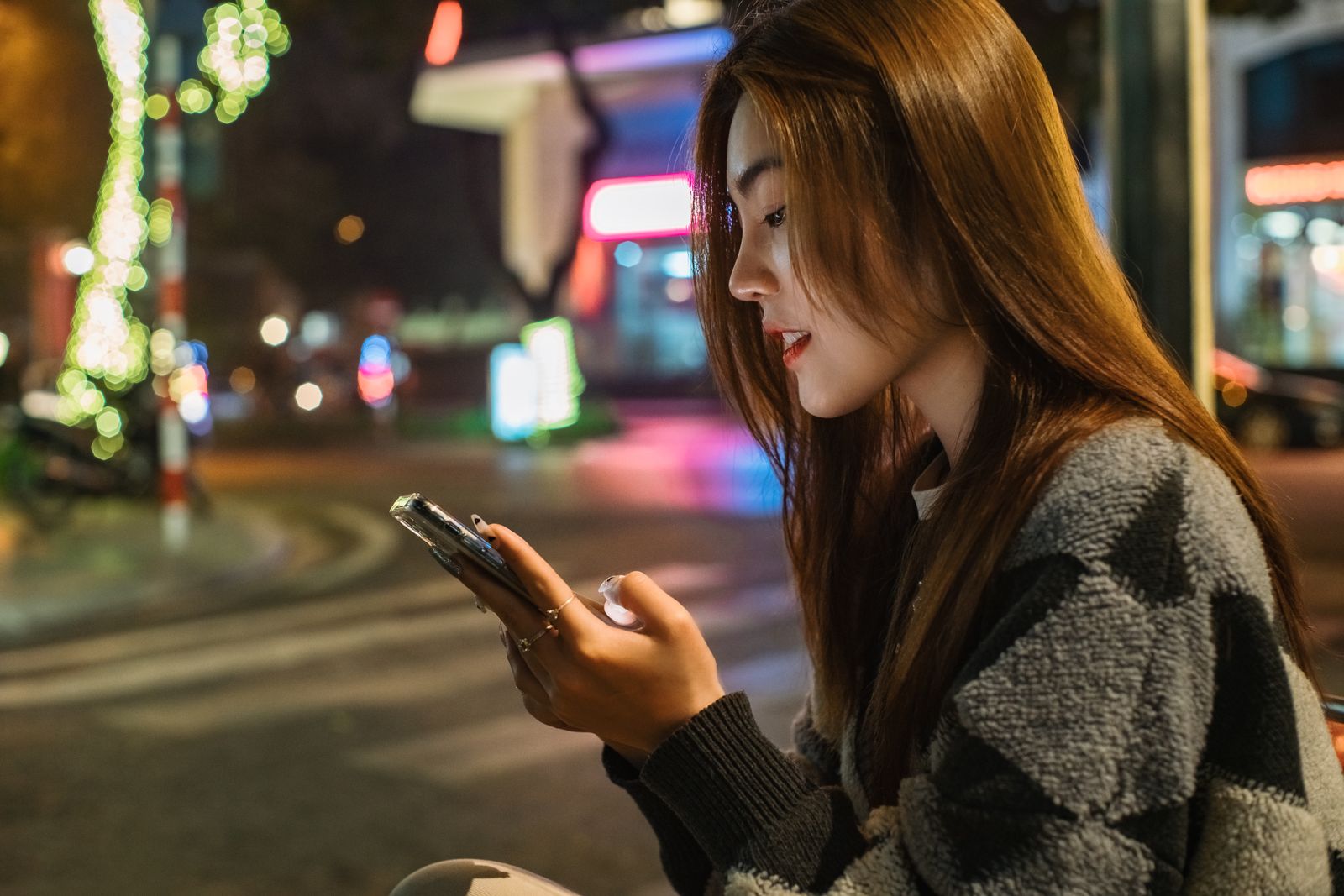 Portrait of a young woman with brown hair using a smartphone on a vibrant urban street at night.