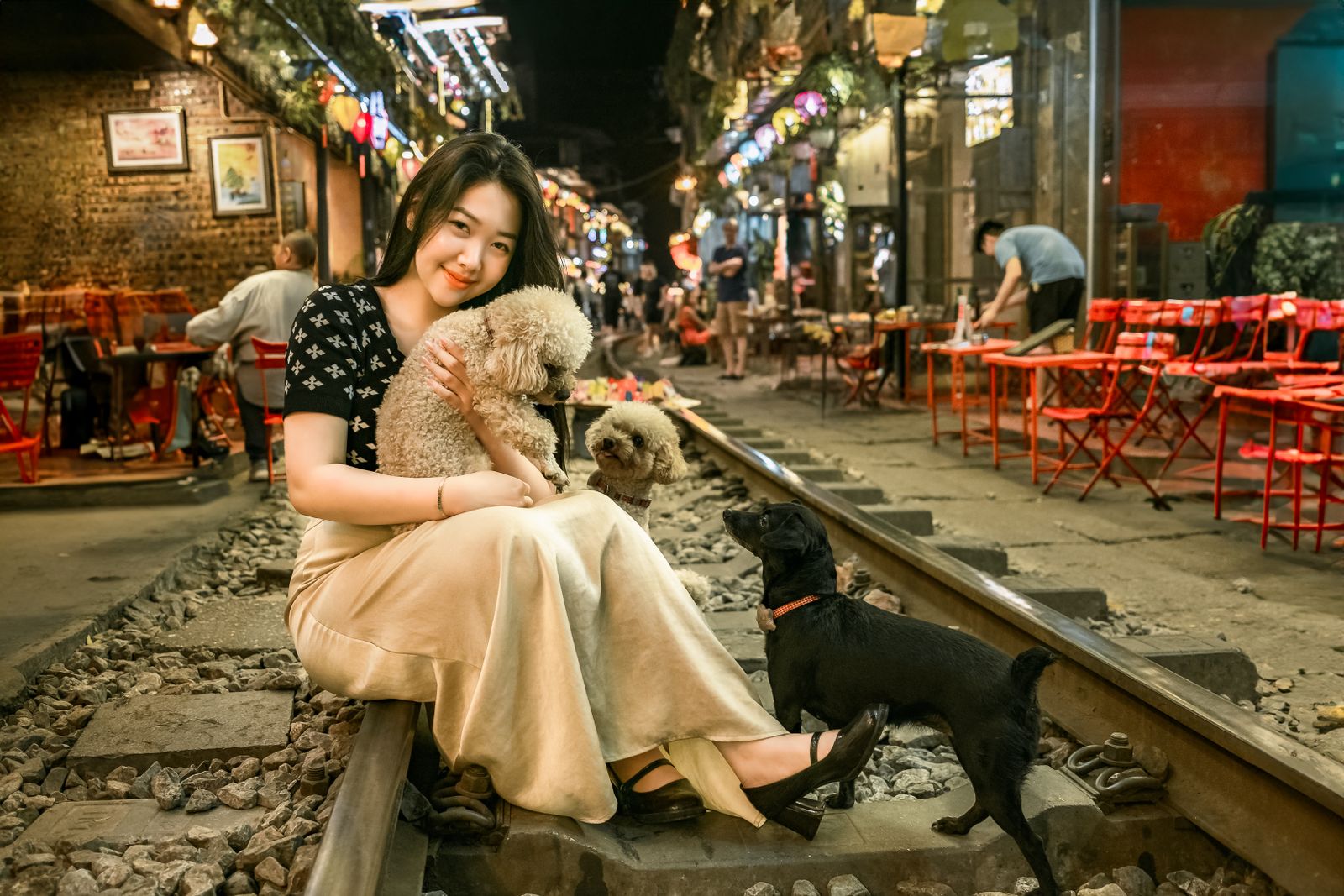 A young woman sitting on Hanoi Train Street with poodles and a black dog, enjoying a vibrant urban evening.