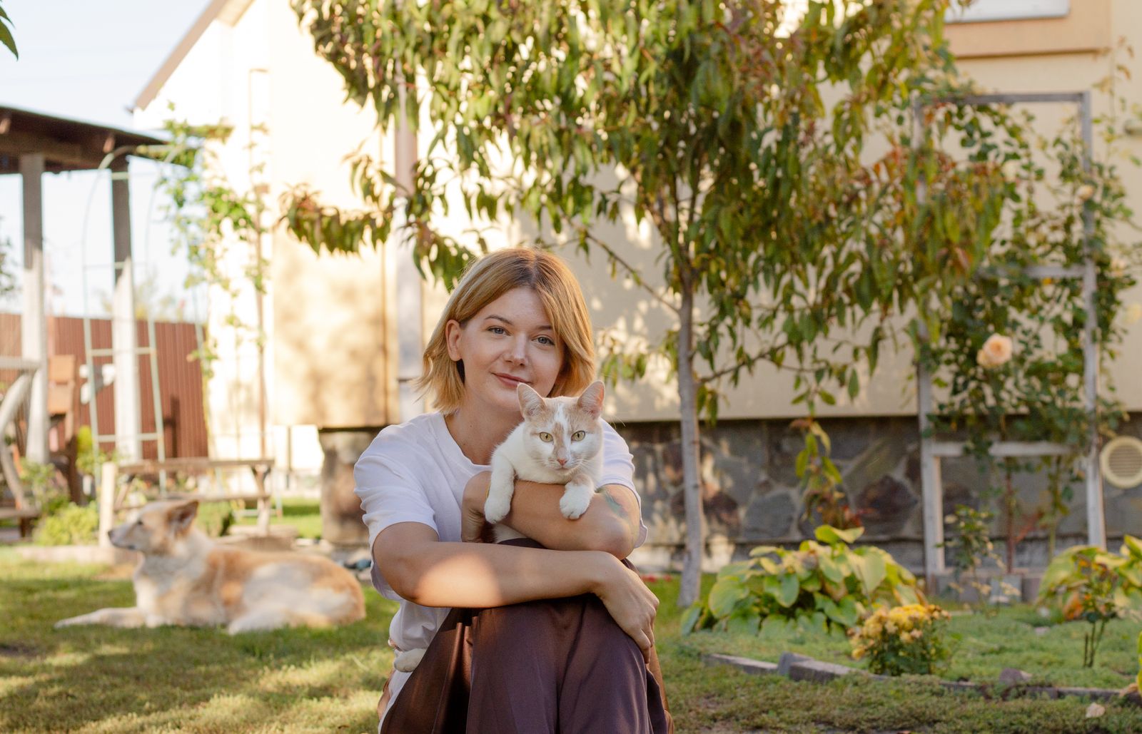 Woman in garden holding a cat, enjoying a sunny day. Relaxed and joyful moment outdoors.