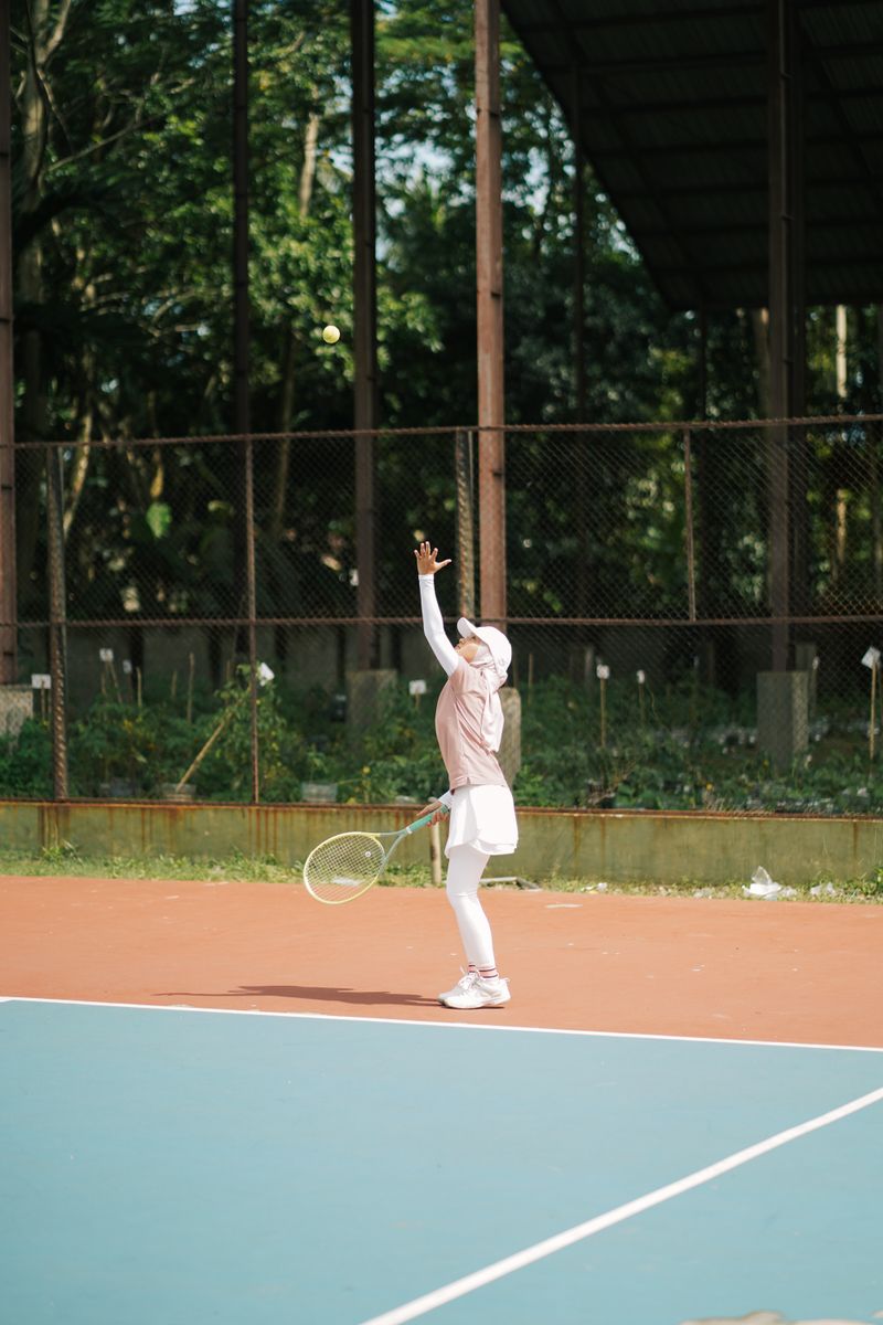 A young woman in sports attire serves on an outdoor tennis court surrounded by greenery.