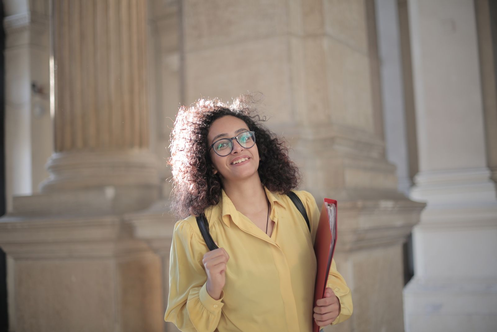 Low angle of gladful woman in round glasses and yellow shirt smiling at camera while standing with folder and backpack on street against blurred exterior of ancient building in old town