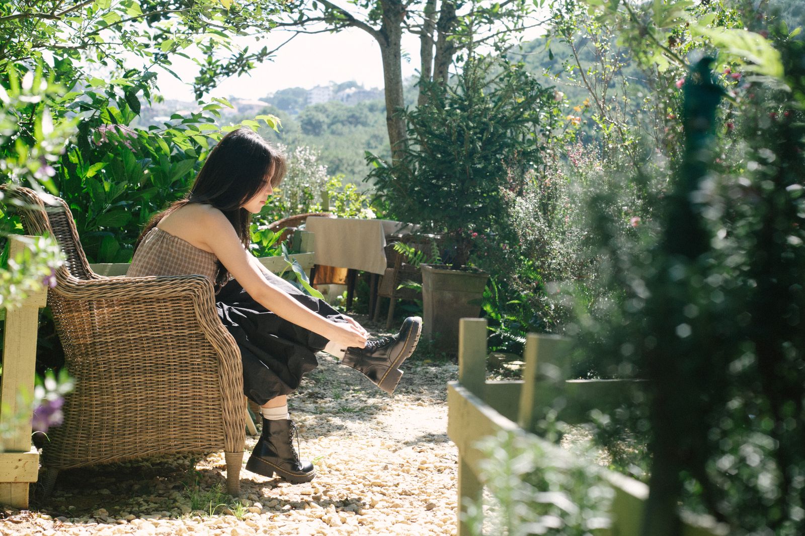 A young woman enjoys the outdoors in a lush garden, sitting on a wicker chair while putting on boots.