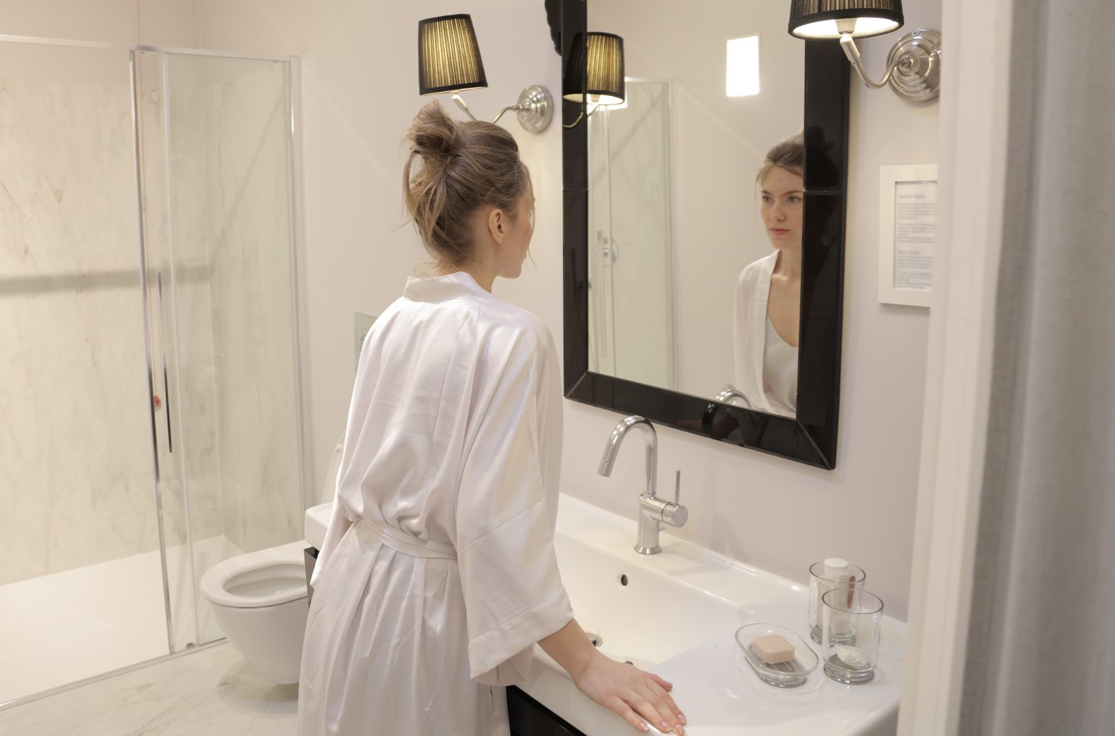 Elegant bathroom scene with a woman in a bathrobe gazing into a large mirror under warm lighting.