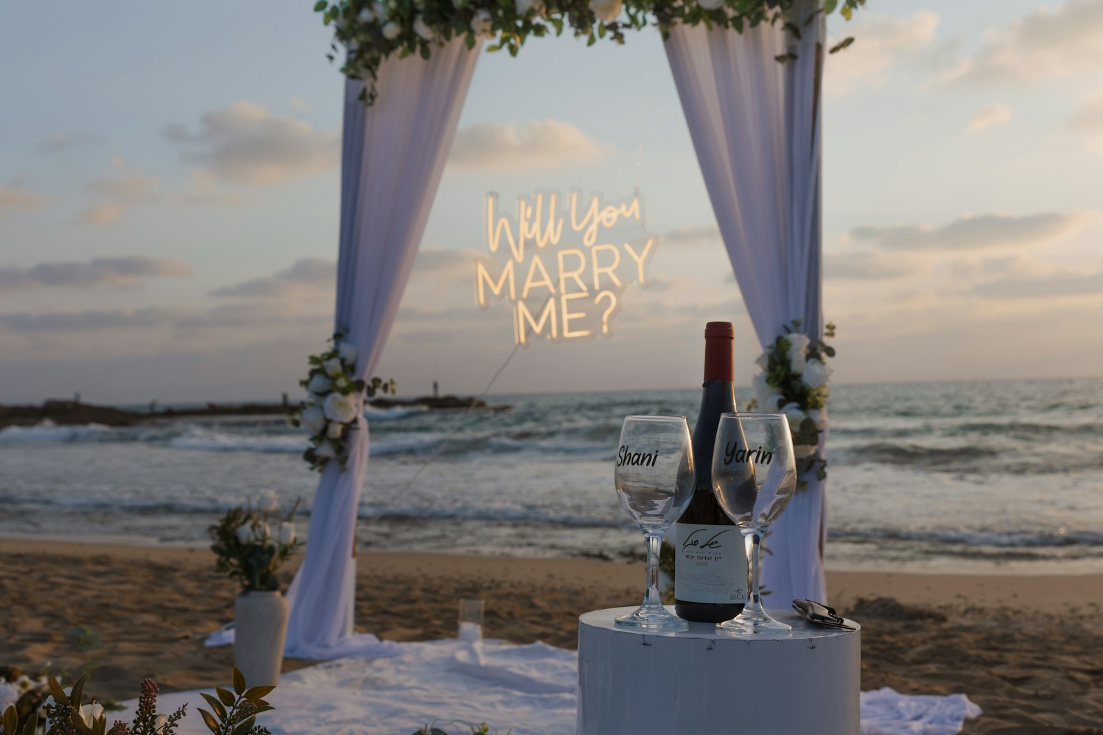 Wedding arch with proposal sign and wine on beach.