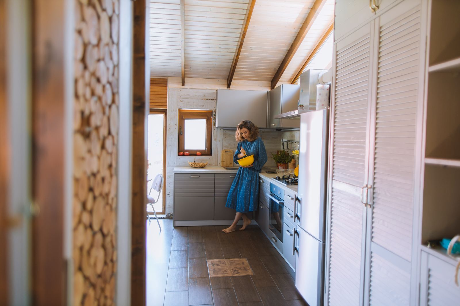 A woman in a blue dress cooking in a cozy, rustic kitchen with modern elements and natural lighting.