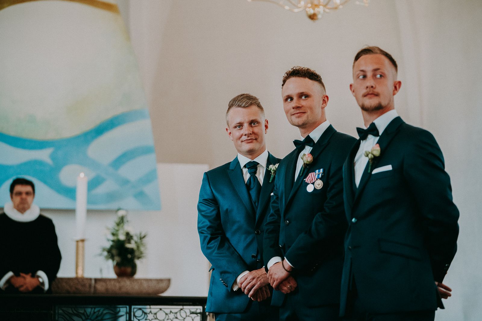 Handsome young groomsmen in elegant black tuxedos decorated with boutonnieres in bright spacious hall