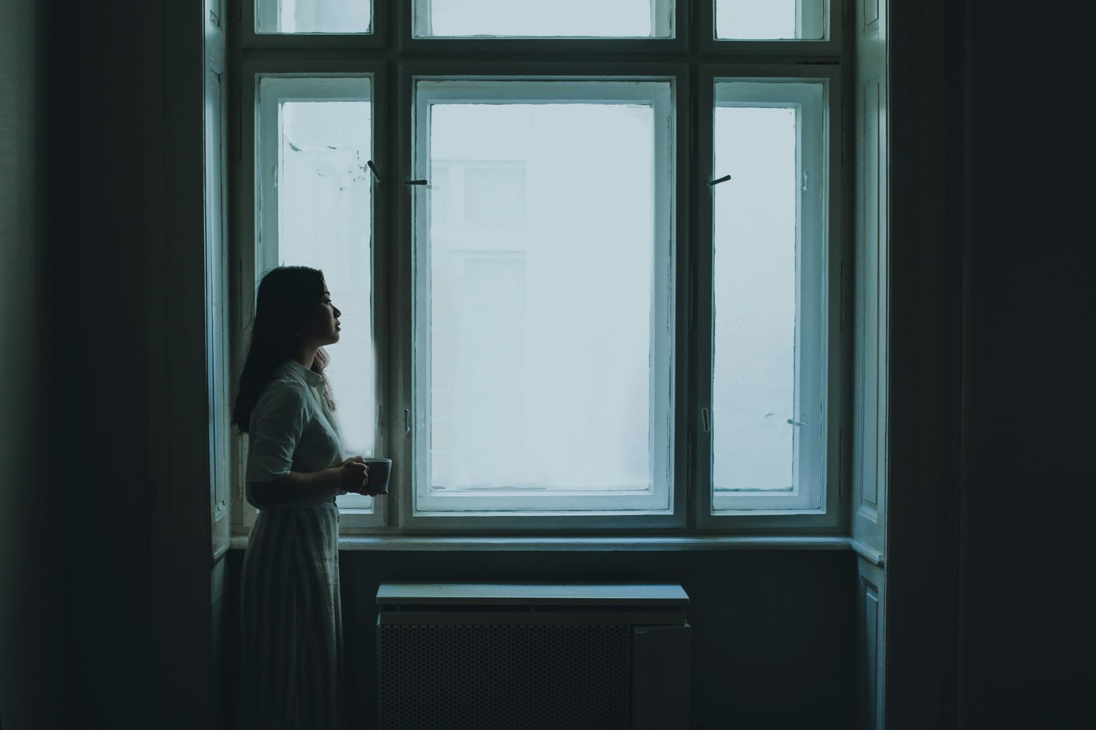Side view of young female in casual clothes standing near window in dark room and drinking coffee in cloudy morning