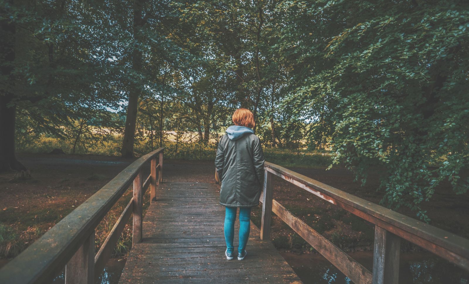 A woman in a jacket walks alone on a wooden bridge in a lush, green forest.
