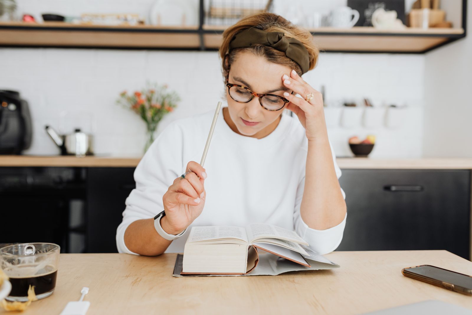 A focused woman in glasses reading a book in a bright, modern kitchen setting with coffee.