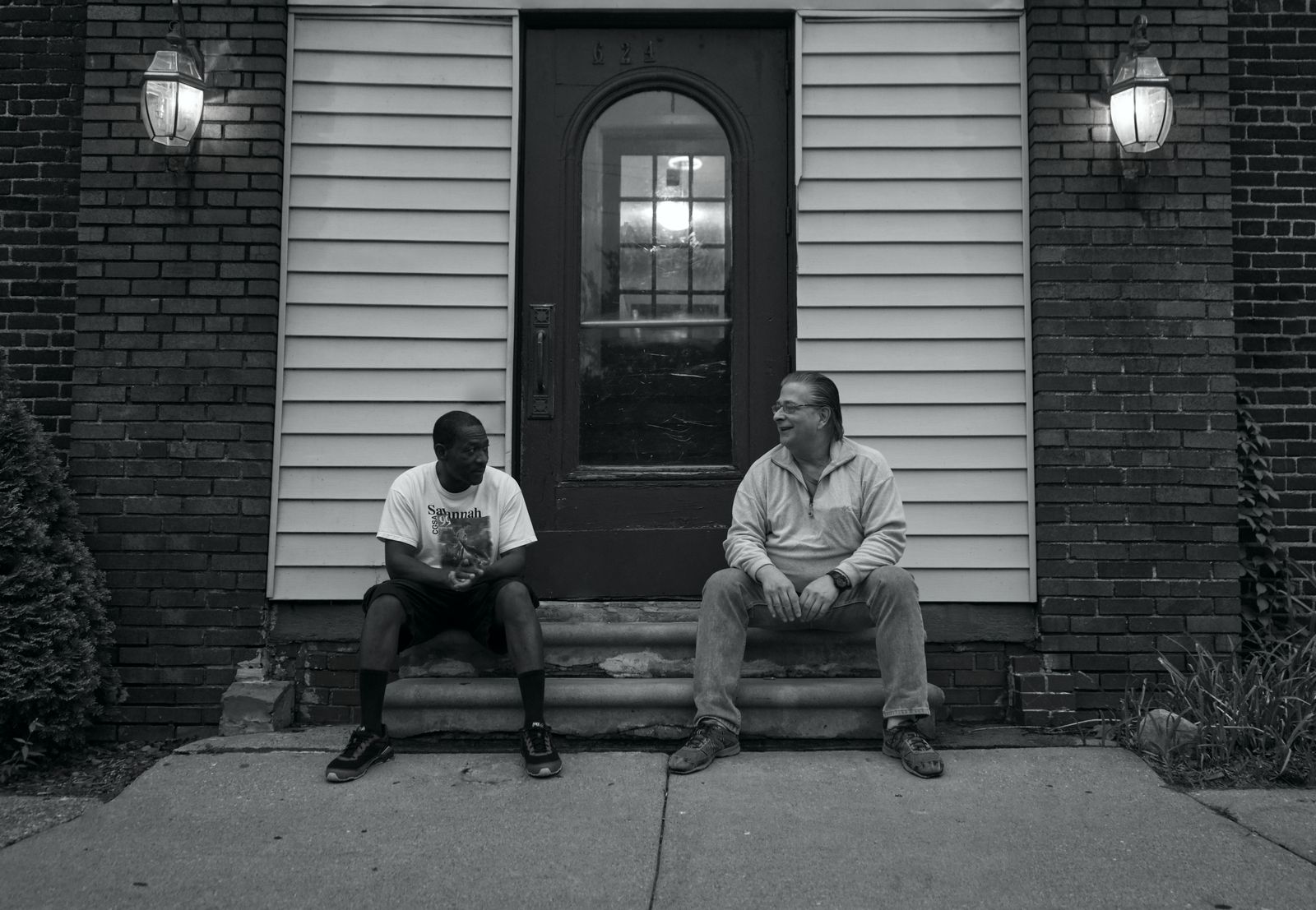 Black and white of multiracial men speaking while resting on concrete stairs of contemporary building with luminous lights in evening