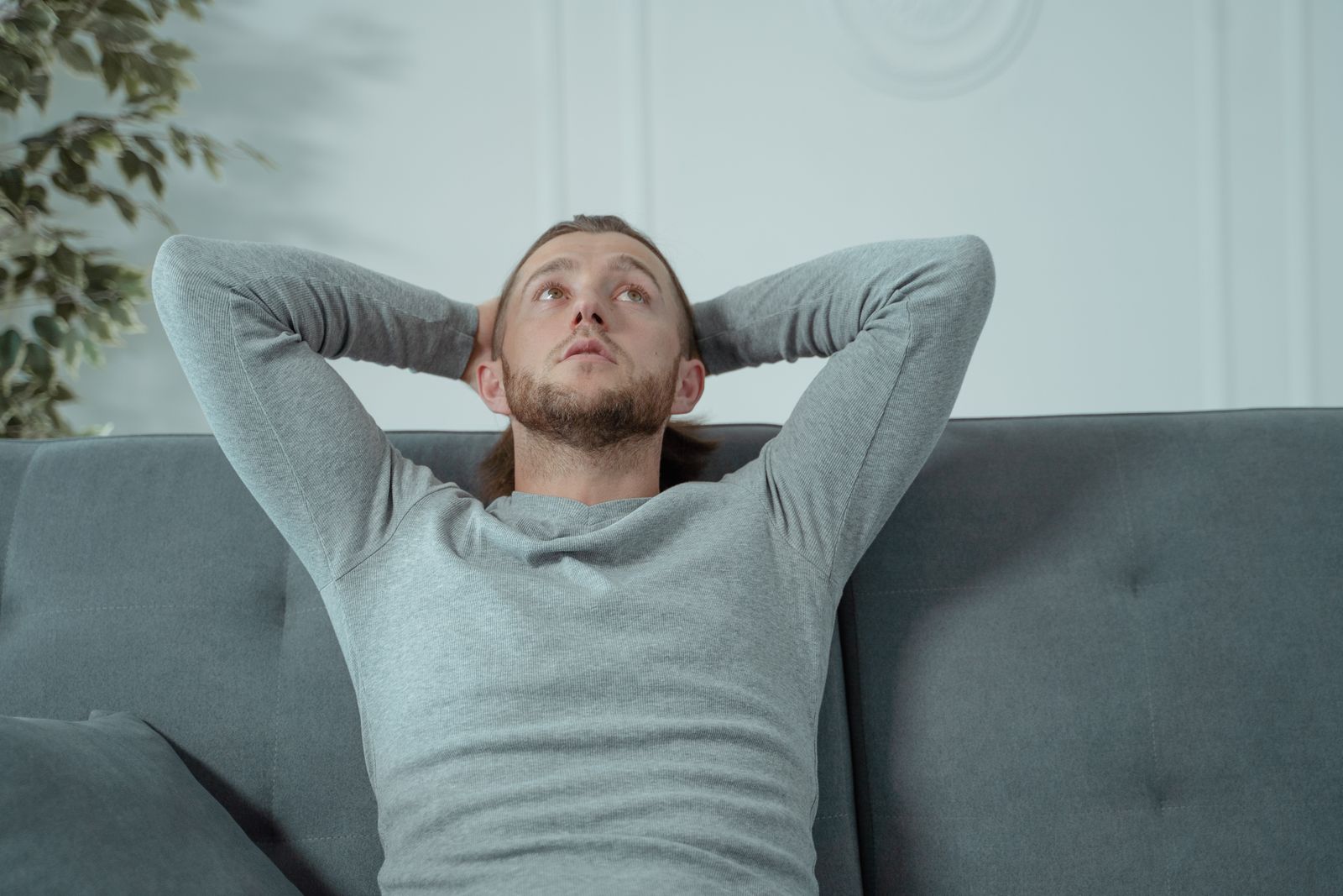 Man looking thoughtful while relaxing on a grey sofa indoors, conveying a sense of contemplation and relaxation.