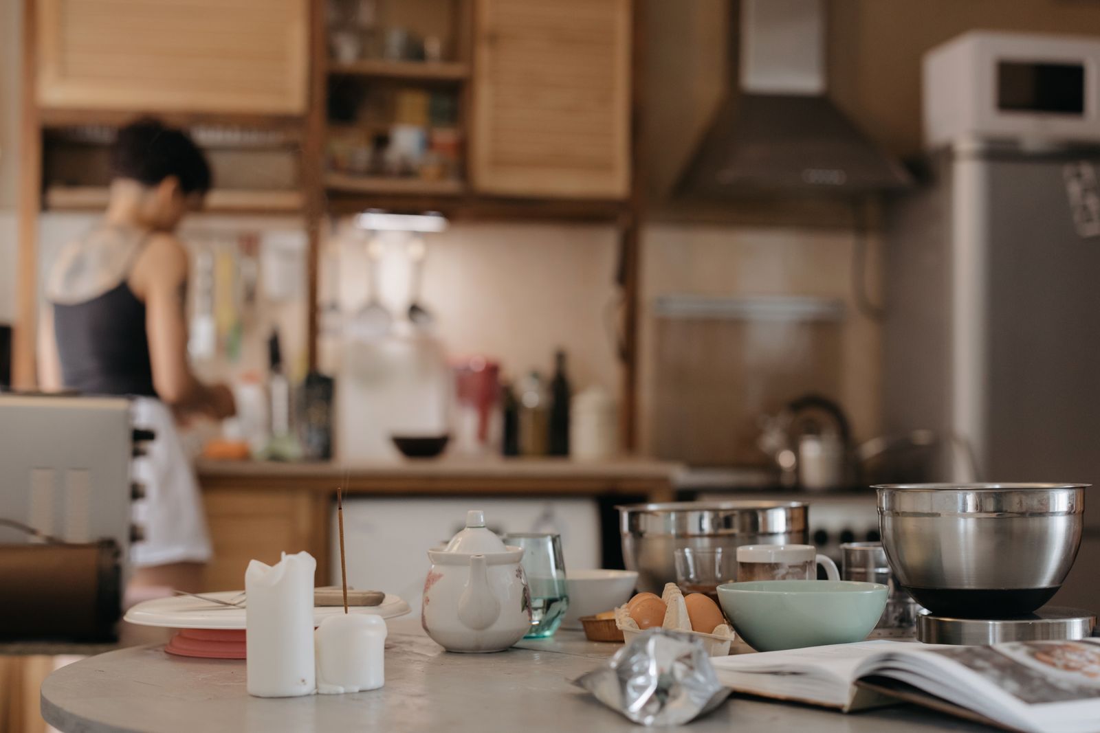 Cozy kitchen scene with a woman cooking, surrounded by utensils and ingredients on the table.