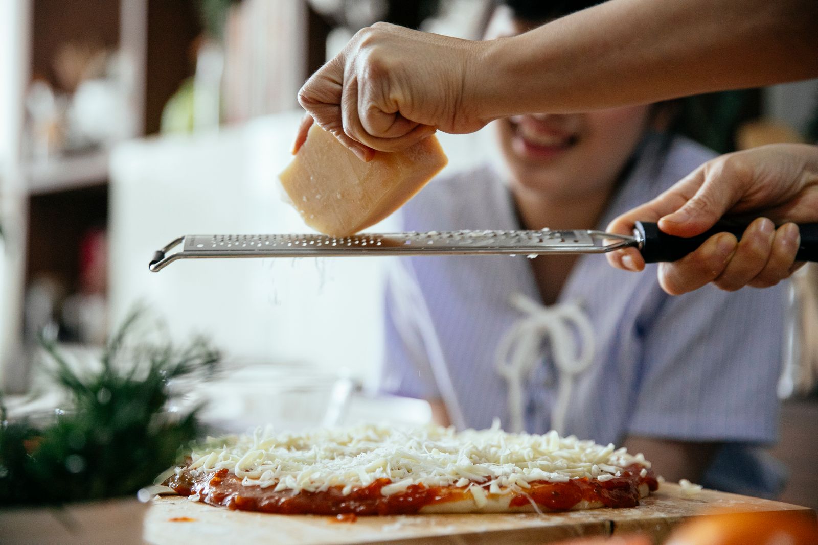 Side view of crop unrecognizable female grating piece of cheese on homemade pizza while cooking in kitchen with friend