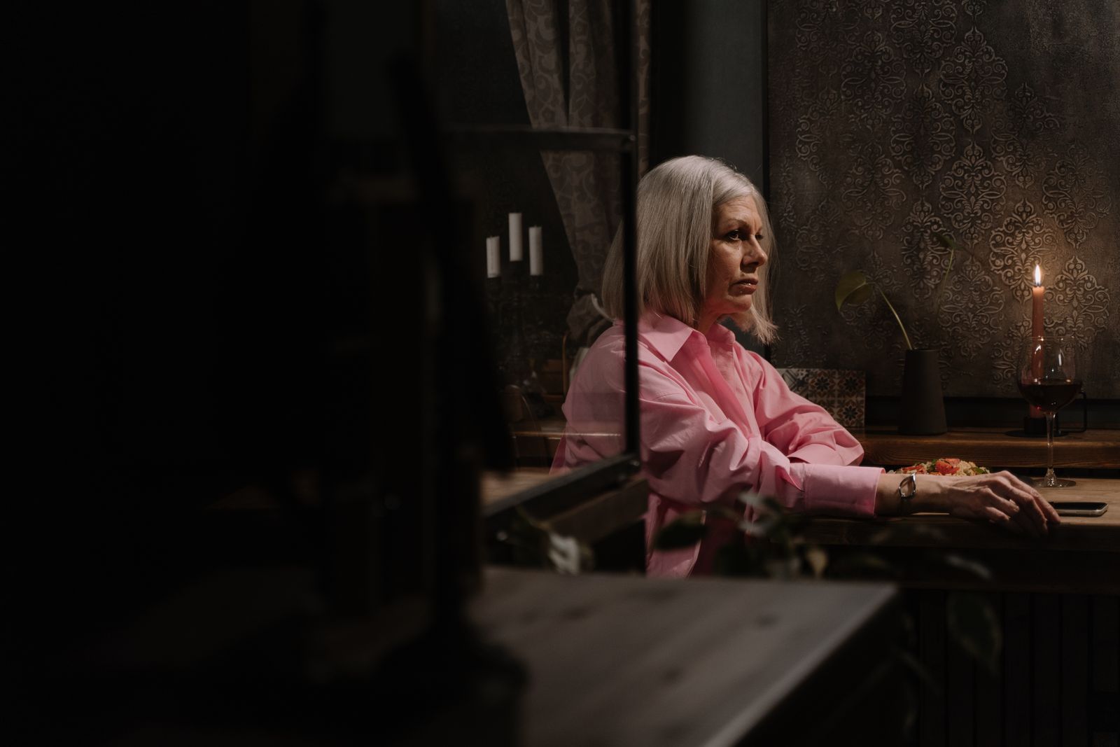 Elderly woman in pink shirt dining alone by candlelight, deep in thought.