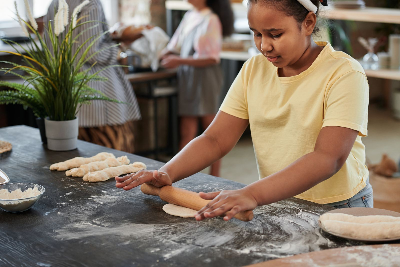A young girl rolls dough with a rolling pin in a bakery setting, surrounded by baking ingredients.