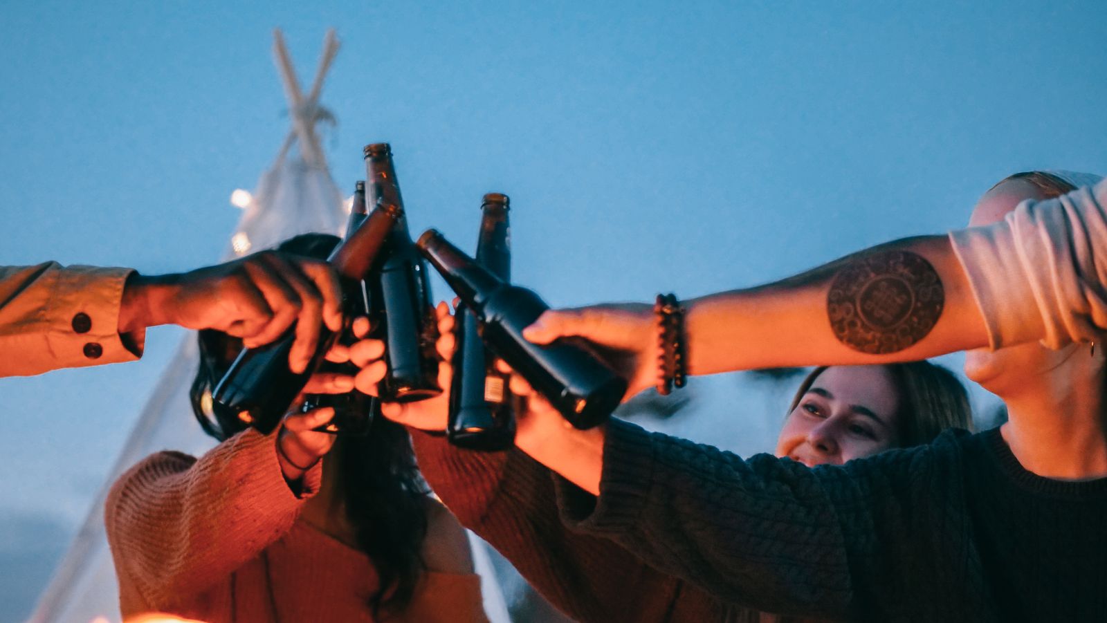 A cheerful group toasting with beer bottles around a beach bonfire at sunset.