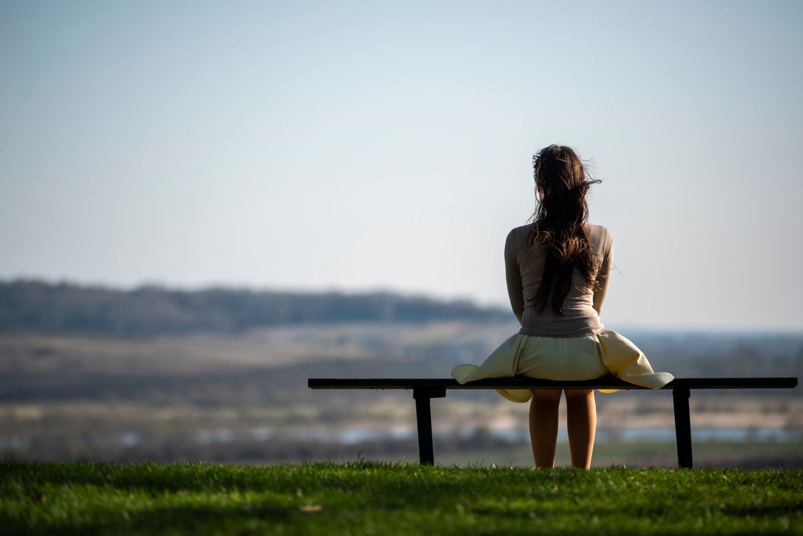 A woman sits on a bench in a field, gazing at the distant horizon under a clear sky.