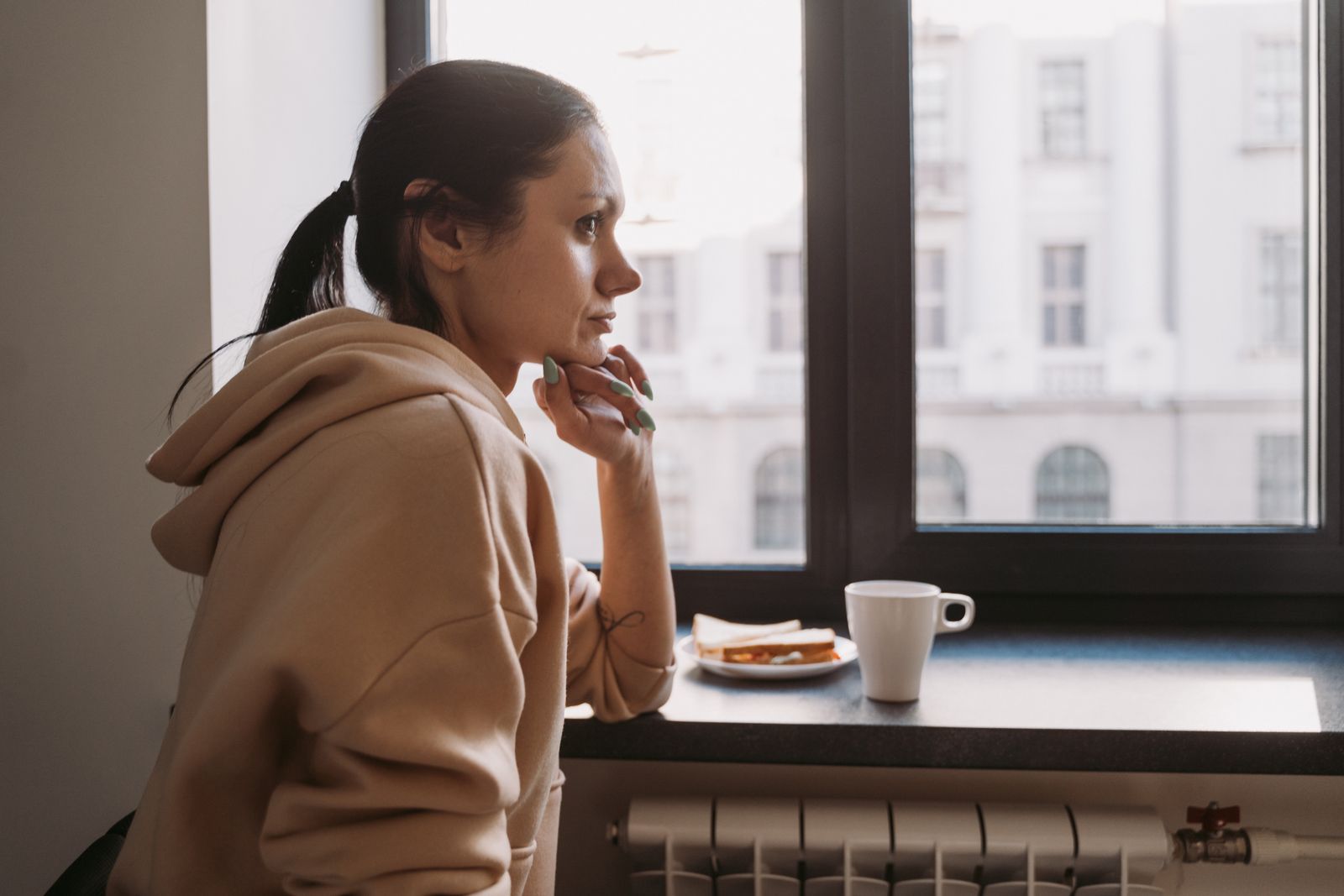 Side view of a woman in a hoodie sitting by a window, contemplating while looking outside.