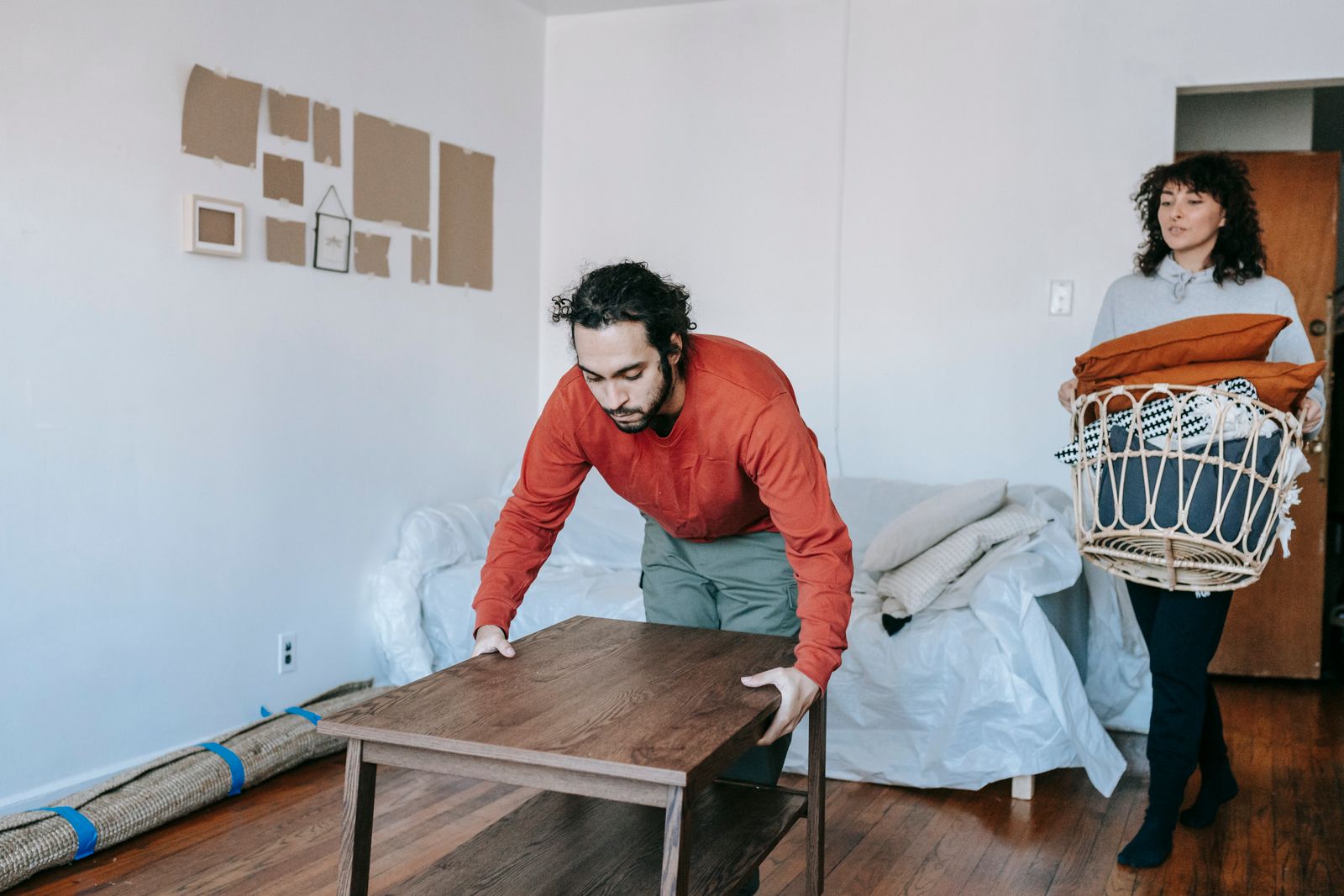 A man adjusts a wooden table while a woman carries a basket in a cozy, contemporary room.