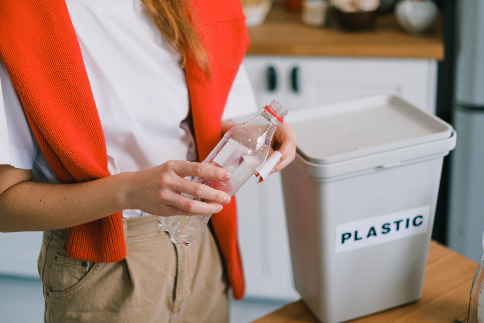 Woman placing plastic bottle into recycling bin in kitchen setting.