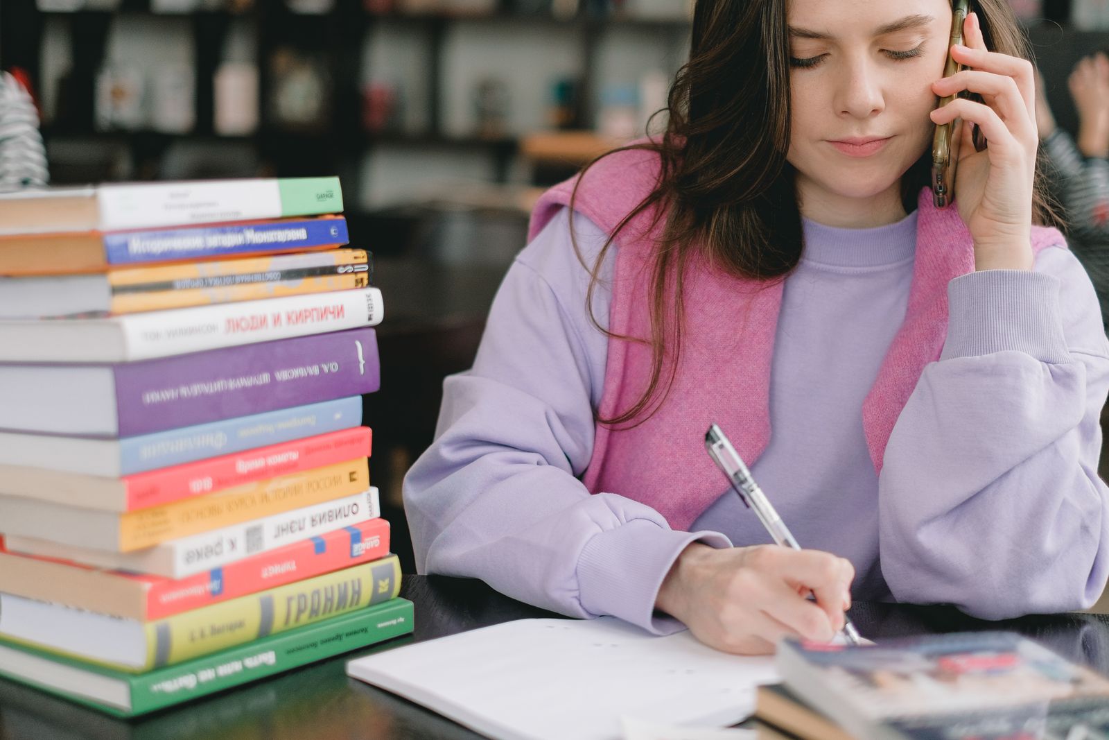 Female student in casual clothes sitting at table with books and writing notes while speaking on phone in university library