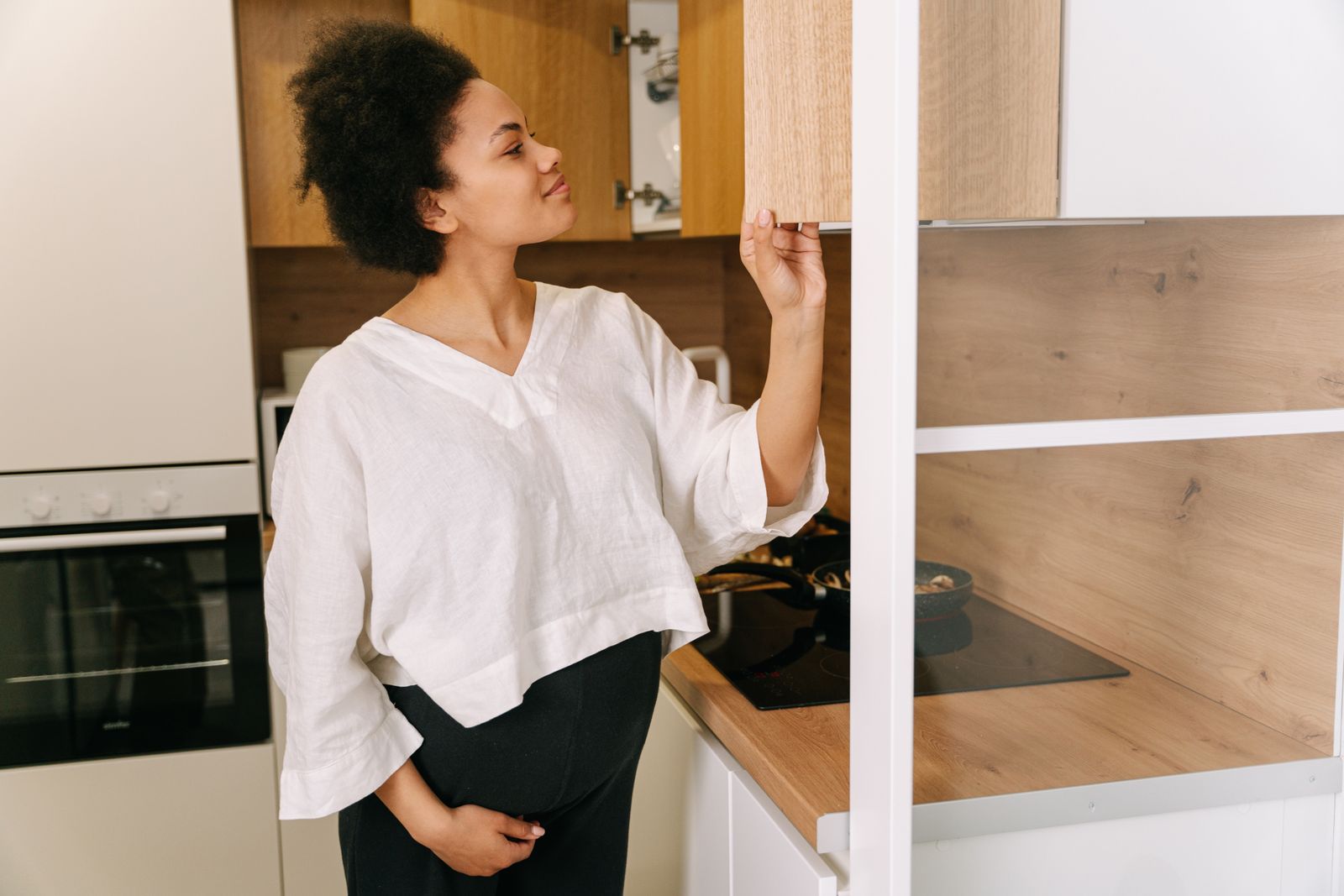 A pregnant woman explores a modern kitchen cabinet while standing near the stove.