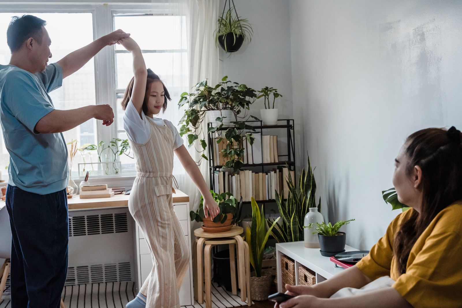 Father dancing with daughter in a sunlit room as mother watches, surrounded by indoor plants.