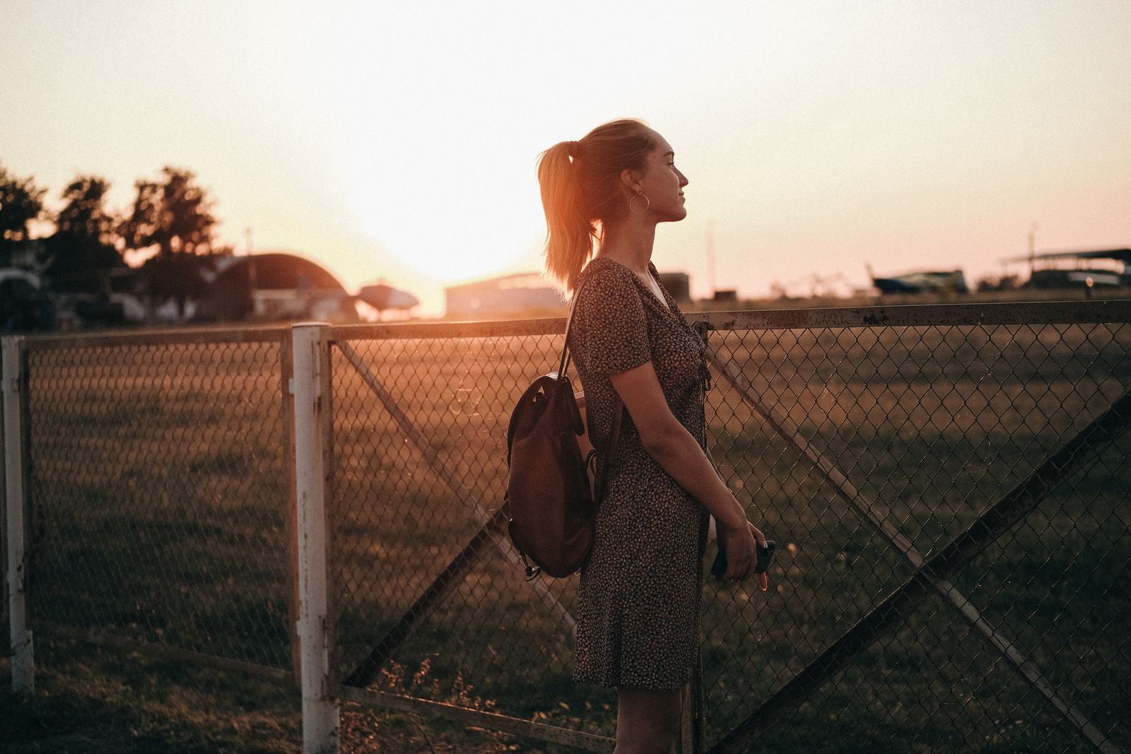 Side view of young dreamy female in dress with backpack looking away against grid fence and airfield in back lit