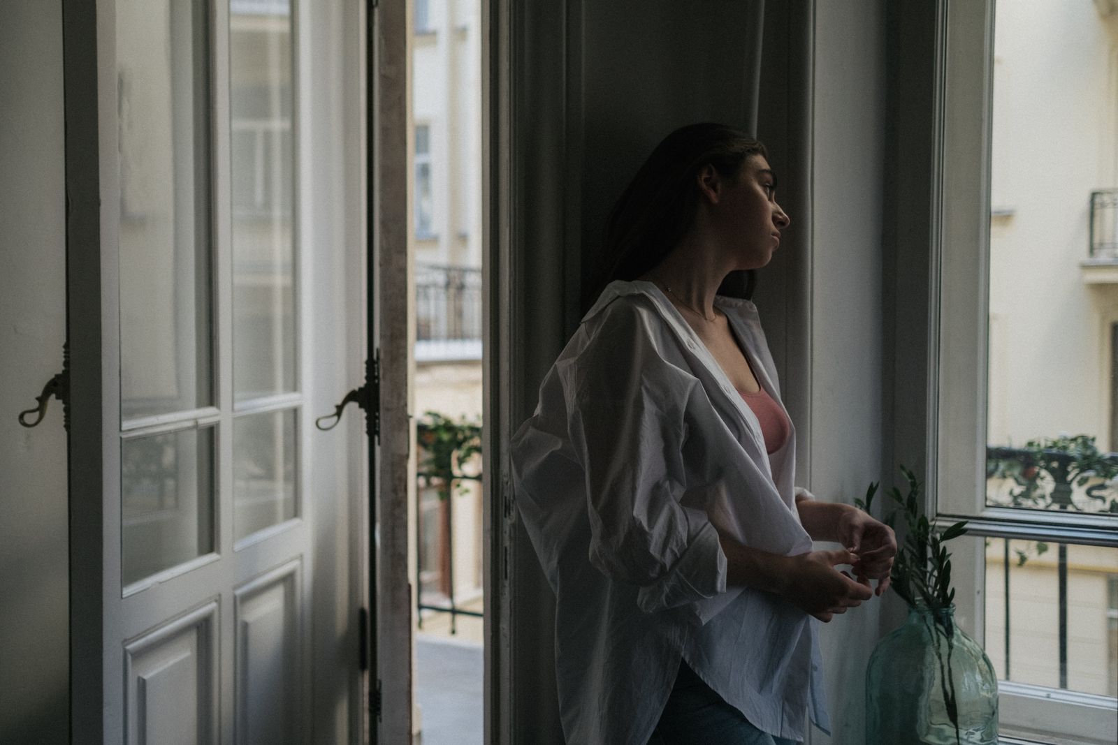 A woman stands by a window, looking outside with a contemplative expression.