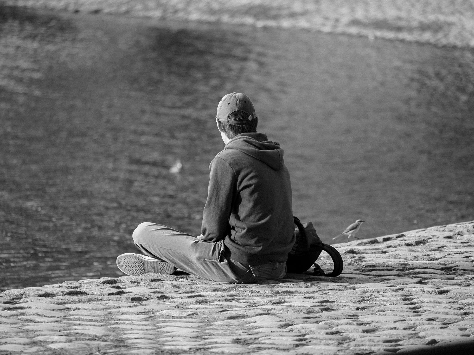 A person in a hoodie sits by a body of water, captured in monochrome tones.