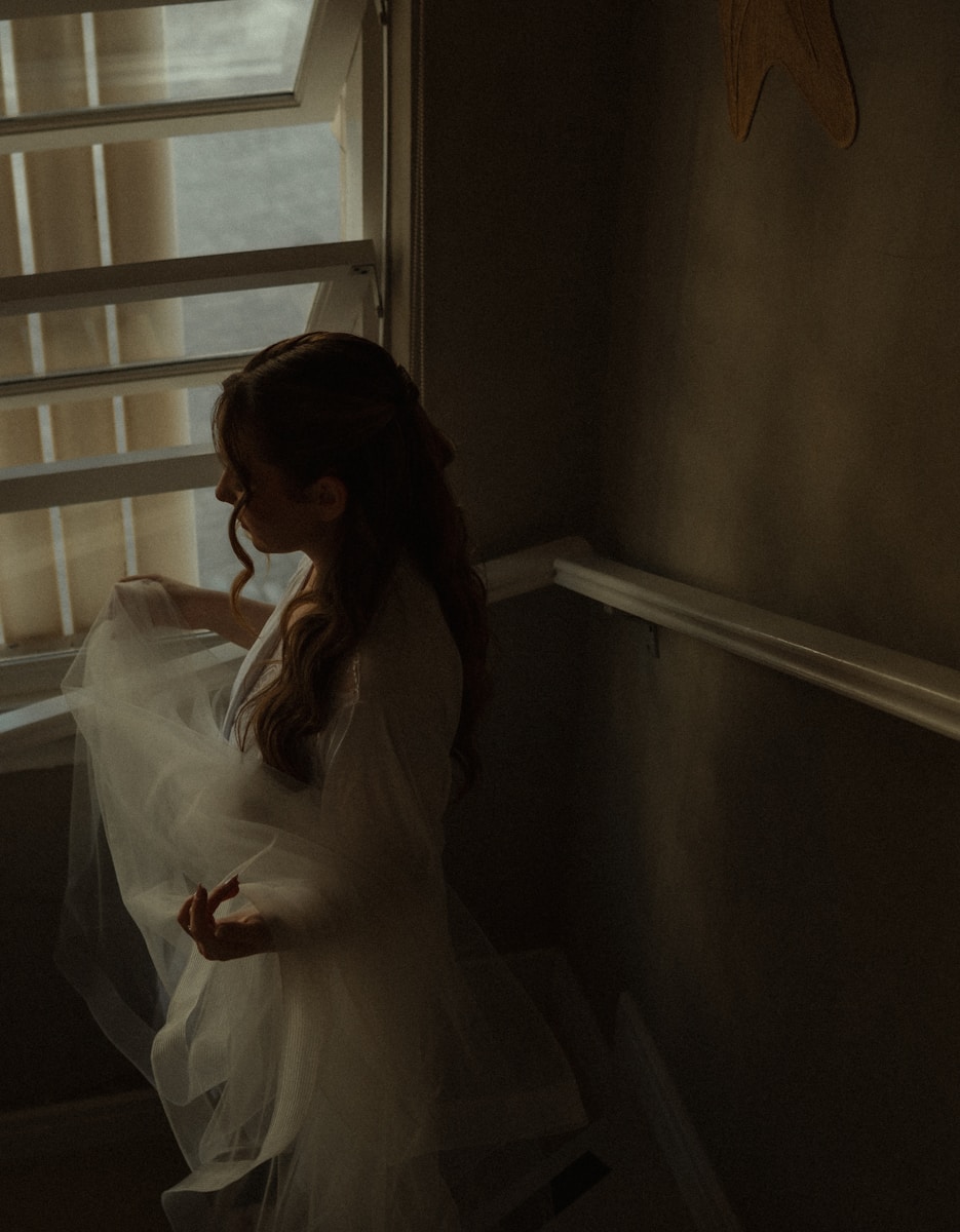 A woman in a sheer dress stands by a window.