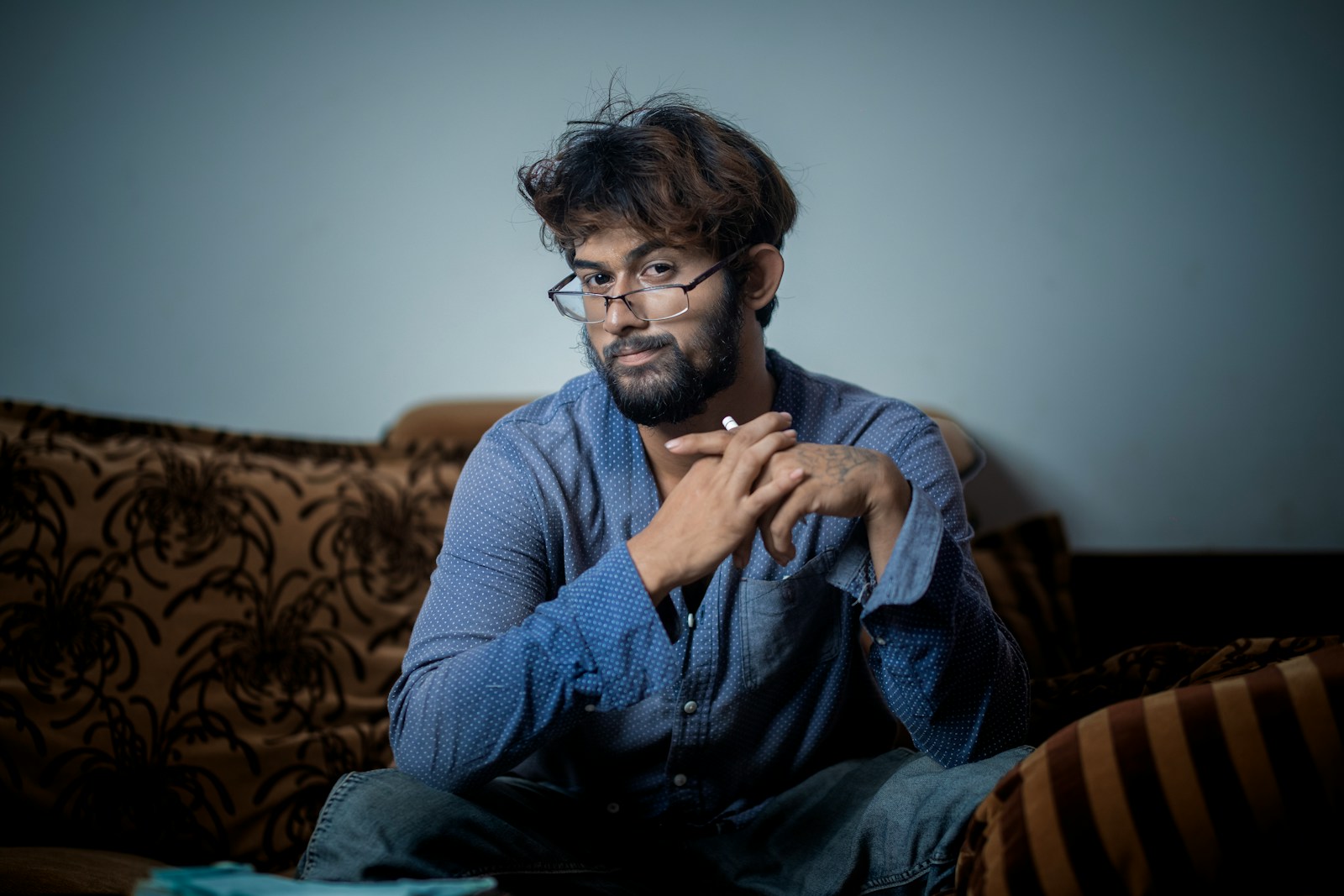 A man with glasses sits thoughtfully on a couch.