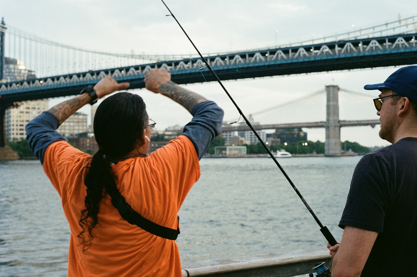Two people fishing near a large bridge over water.