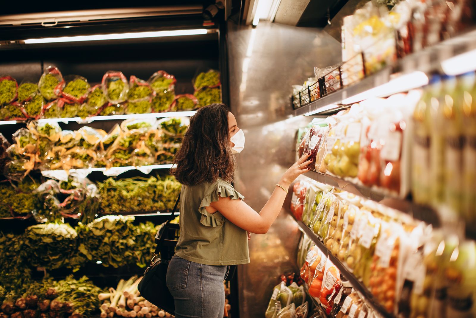Woman wearing a mask choosing fresh produce in a supermarket aisle.